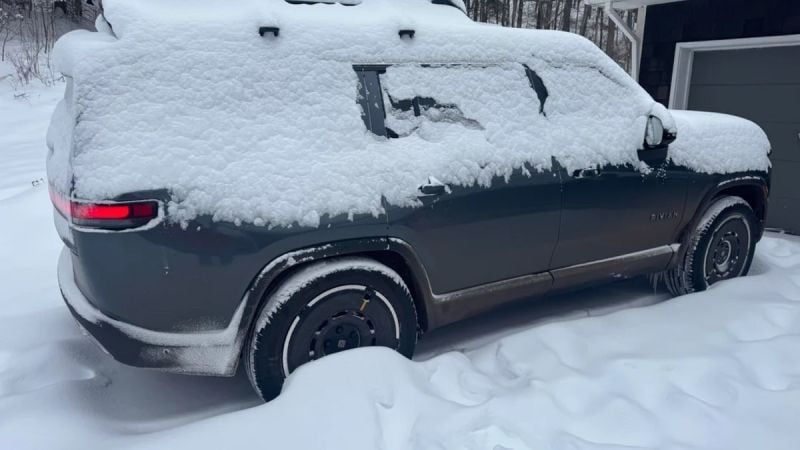 A dark-colored Rivian electric vehicle covered in heavy snow, showing the rear quarter view with illuminated taillights and snow-covered wheels in deep winter conditions.