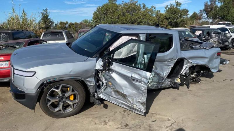 A severely damaged silver Rivian R1T, showing extensive side impact damage and crumpling along its passenger side while parked in what appears to be a salvage yard.