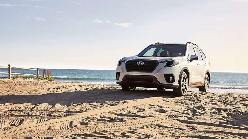 A Subaru SUV parked on sandy beach shore, with ocean waves crashing gently in the background under a clear sky.