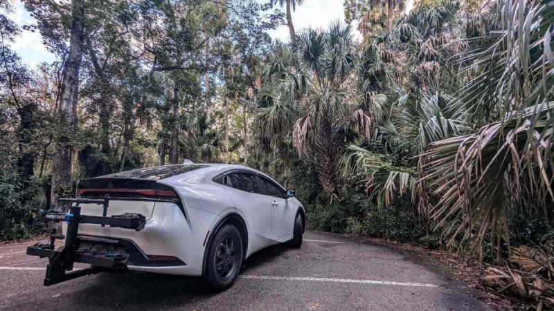 2023 white Toyota Prius hybrid with rear bike rack parked in a forest, rear three-quarter view.