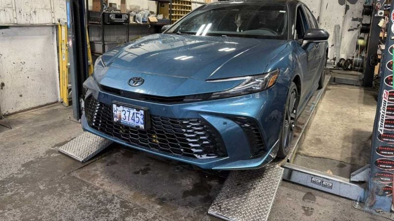 2023 Toyota Camryin Guard Metallic on a hydraulic lift in a repair shop, front three-quarter view showing the full car body and surrounding tools.