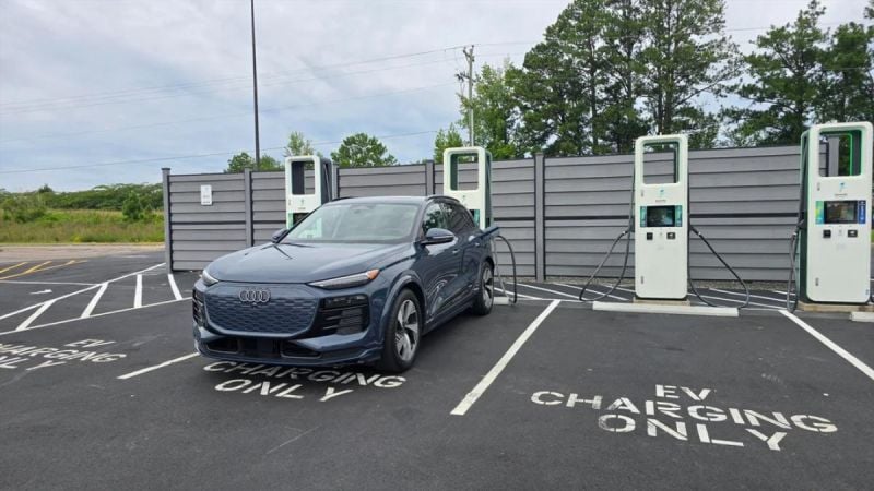 Dark blue 2024 Audi Q6 e-tron SUV plugged in at EV charging station with gray privacy wall backdrop