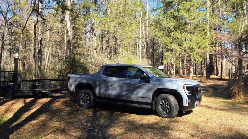 A silver 2024 GMC Sierra EV pickup truck shown from a front 3/4 angle, parked in a wooded area with tall pine trees in the background.