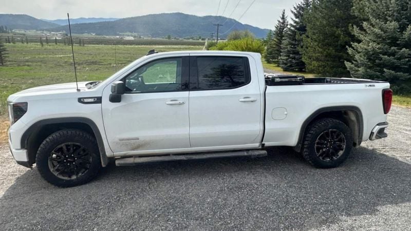 White 2024 GMC Sierra pickup truck with black wheels and off-road tires, photographed from side angle in mountainous rural setting with evergreen trees.