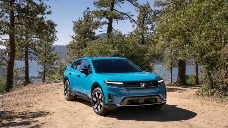 Bright teal 2024 Honda Prologue Elite SUV parked on dirt path with pine trees and lake in background