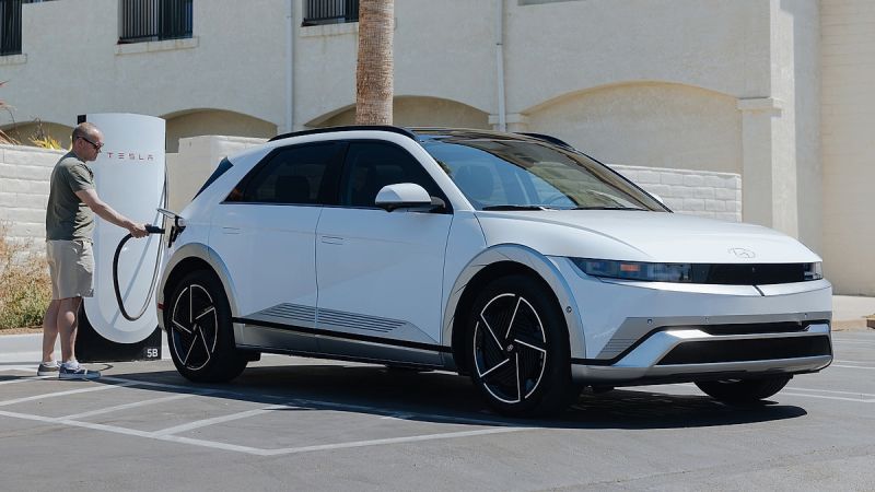 2024 Hyundai Ioniq 5 in white parked at a Tesla Supercharger station with an owner plugging in the NACS charging cable demonstrating the Ioniq 5s expanded charging network access in a sunny suburban parking lot