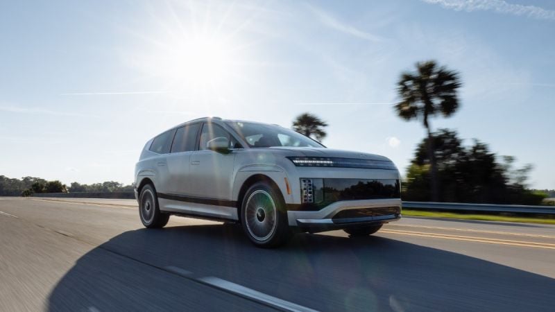 2024 Hyundai IONIQ 9 electric SUV in silver, shown from 3/4 front angle driving on coastal road with palm trees, featuring distinctive LED lighting and aerodynamic wheels.