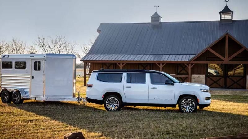 A white 2024 Jeep Wagoneer towing a silver horse trailer near a rustic barn on grassy land, illuminated by warm sunlight.