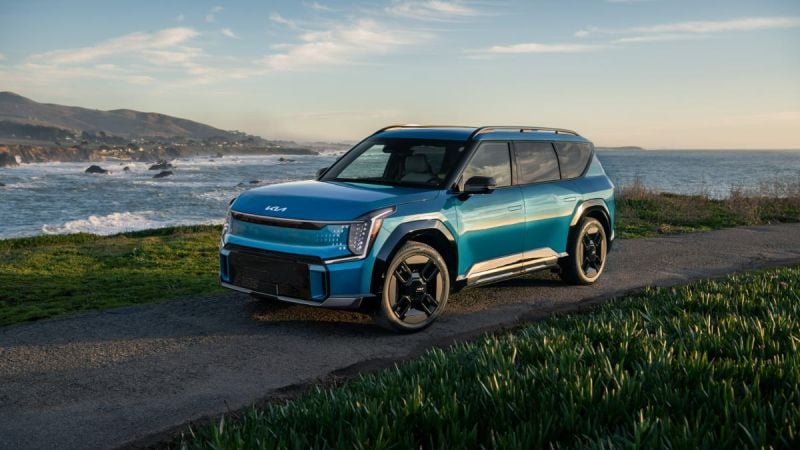 A teal blue 2024 Kia EV9 electric SUV is shown from a front three-quarter angle, parked on a coastal overlook with dramatic ocean waves and mountains in the background.