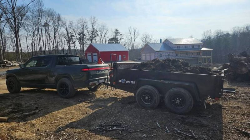 Pickup truck towing a trailer filled with soil, near red and white buildings, surrounded by trees.