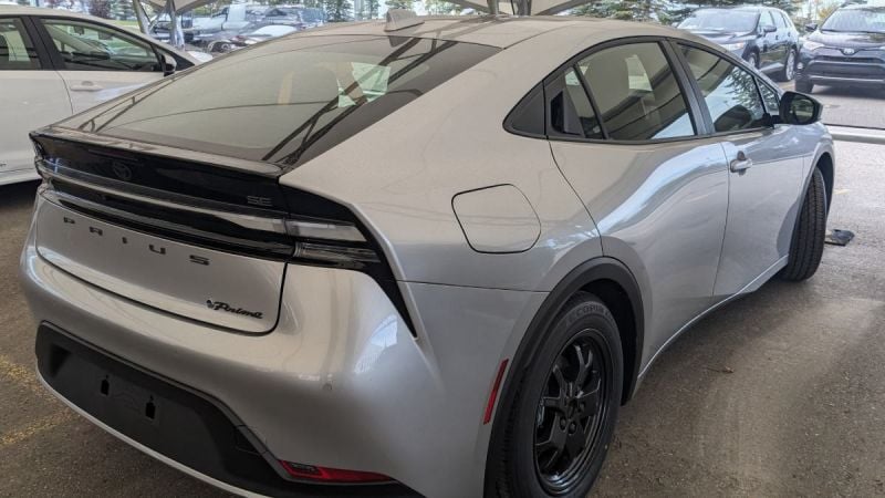 2024 Toyota Prius Prime in silver, rear three-quarter view, featuring black alloy wheels and distinctive fastback design, parked under dealership canopy.