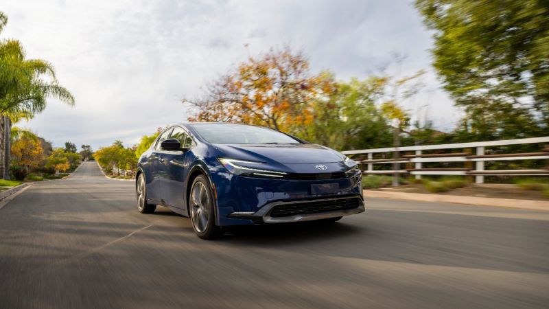 A navy blue 2024 Toyota Prius shown from a front three-quarter angle, driving on a tree-lined suburban road with fall foliage in the background.