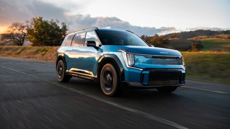 Blue 2024 Kia EV9 electric SUV parked on a mountain road with green hills and dramatic clouds in the background.