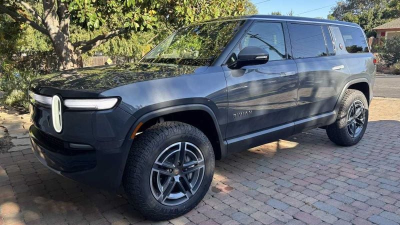 2024 Rivian R1S in dark gray parked on a brick driveway with a large tree and suburban house in the background.