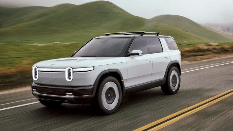 Silver Rivian R2 electric SUV driving on a paved road through rolling green hills with motion blur emphasizing speed.