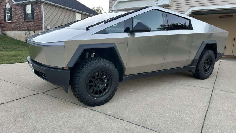 Side view of a Tesla Cybertruck in metallic silver, showcasing its angular design and black off-road wheels with aggressive all-terrain tires parked on a concrete driveway.