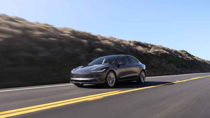 2024 Tesla Model 3 in Midnight Silver Metallic driving on a coastal highway, front three-quarter view with hillside and blue sky background