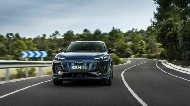 2025 Audi Q6 e-tron in blue, front view on a winding road, showcasing distinctive grille and headlights.
