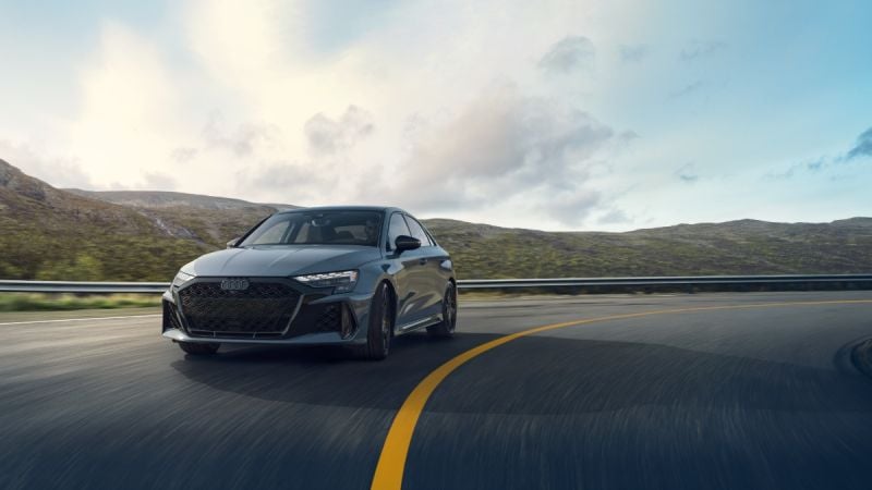 2025 Audi RS3 in dark gray, front three-quarter view, driving on mountain road with aggressive black grille and sleek LED headlights.