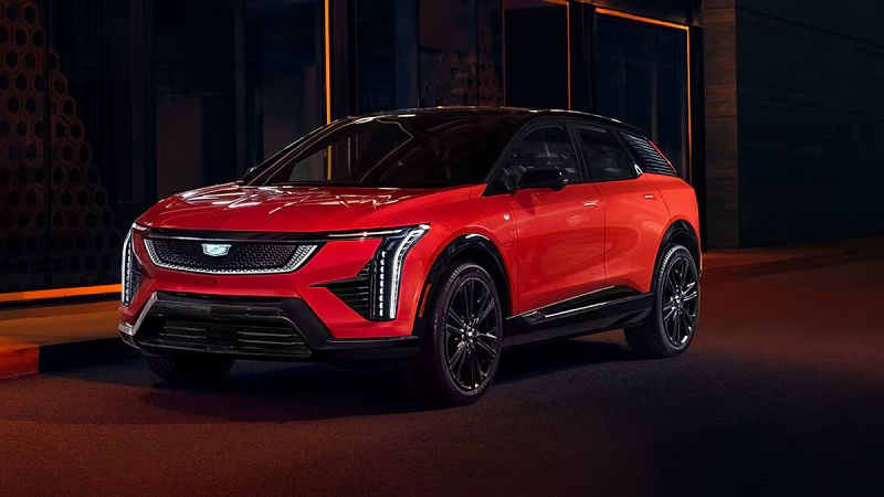 2025 Cadillac Optiq Sport in infrared red with a gloss black roof posed under dramatic nighttime lighting with sharp angular LED headlights glowing highlighting the luxury electric crossover front fascia design