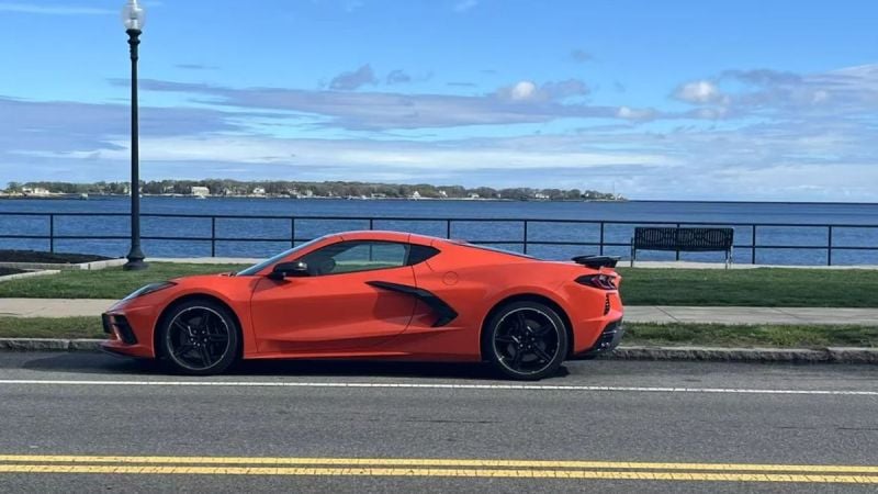 Bright orange 2025 Chevrolet Corvette C8 parked oceanside, side profile view, featuring black wheels and aggressive styling, against scenic coastal backdrop.