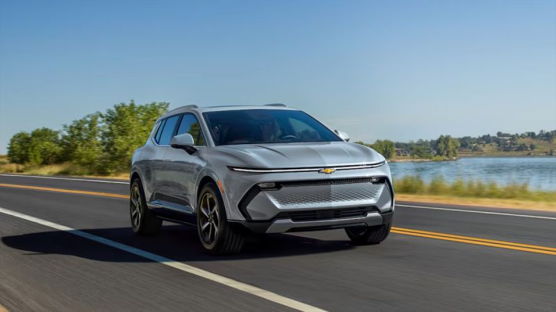 Silver Chevrolet Equinox EV SUV driving on scenic lakeside highway with trees and blue sky