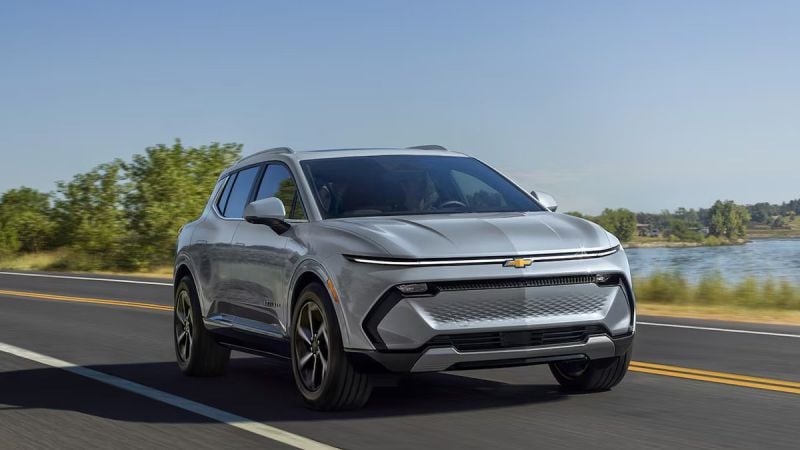 A silver Chevrolet SUV driving on a scenic road with greenery and a body of water in the background under a clear blue sky.