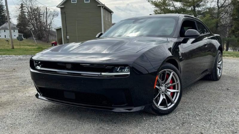 A 2025 Dodge Charger EV with red brake calipers parked on gravel, surrounded by green grass and trees in the background.