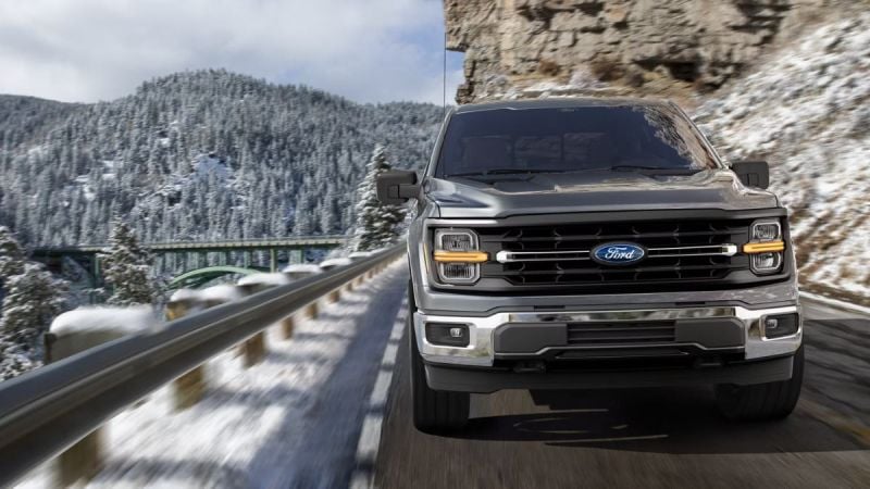 A silver 2025 Ford F-Series pickup truck shown from the front quarter angle, driving on a mountain road with snow-covered peaks in the background.