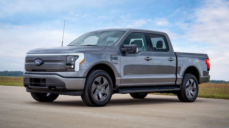2022 Ford F-150 Lightning electric pickup truck in silver, three-quarter front view, featuring distinctive LED light bar and closed grille, parked on concrete with rural landscape background.