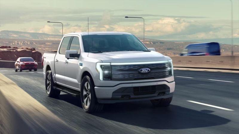 White 2025 Ford F-150 Lightning electric pickup truck driving on highway with mountain landscape in background