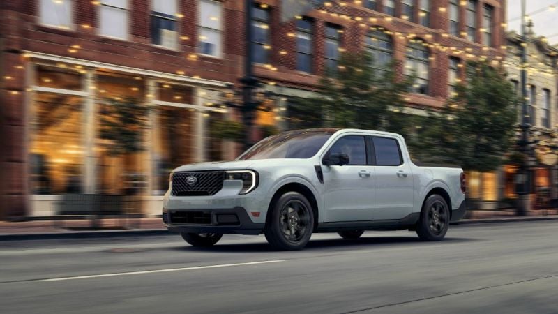 A white 2023 Ford Maverick pickup truck shown from a front three-quarter angle, driving on a city street with motion blur and brick buildings in the background.