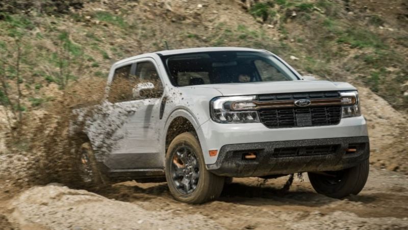 White pickup truck driving through muddy terrain, splashing mud around, with grassy background.
