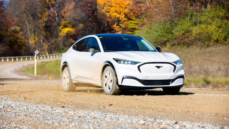 White Ford Mustang Mach-E electric SUV driving on gravel road with autumn forest background