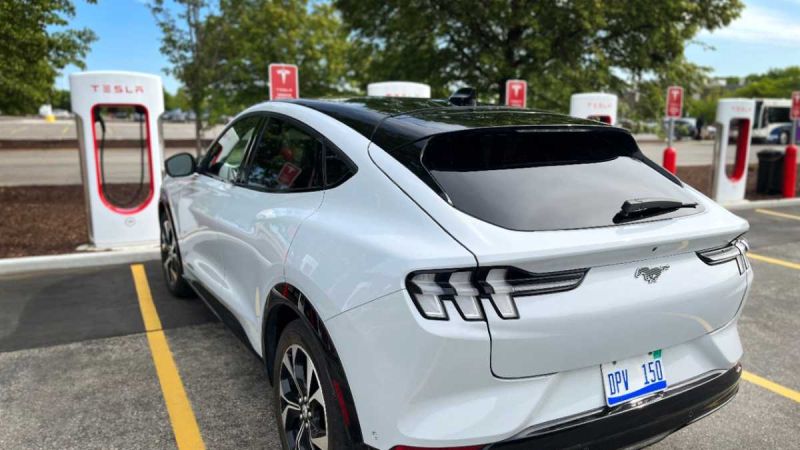 A white Ford Mustang Mach-E parked at a Tesla charging station, surrounded by trees and a clear blue sky.