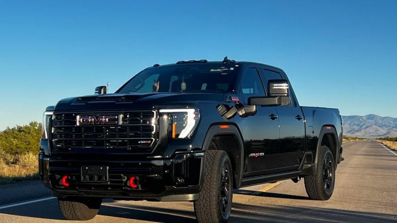 Black 2024 GMC Sierra HD pickup truck photographed from front quarter angle, featuring bold chrome grille, lifted suspension, and off-road tires, parked on desert highway with mountains behind.