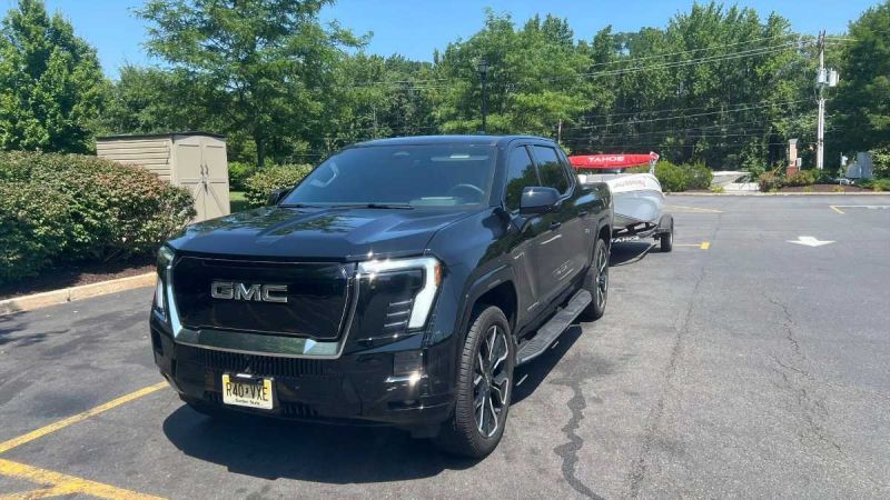 Dark blue 2025 GMC Sierra EV pickup truck parked in suburban lot with trees and utility pole in background, angled front three-quarter view showcasing bold grille and unique design.