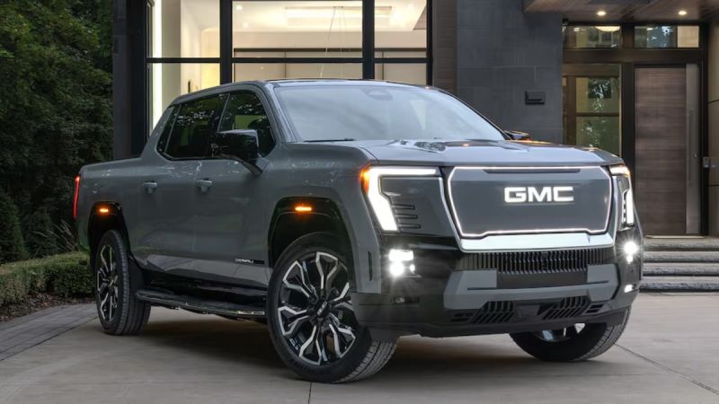 Dark gray 2025 GMC Sierra EV Denali pickup truck parked in front of modern glass and stone building with LED headlights on