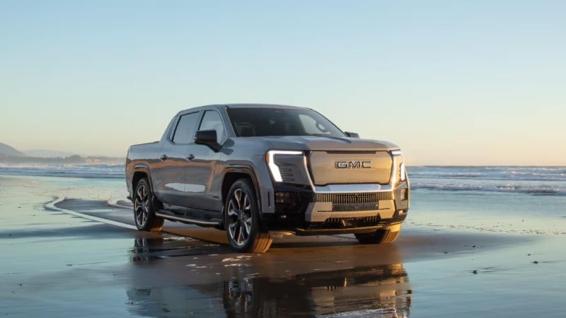 2025 GMC Sierra EV Denali pickup truck in silver parked on a reflective beach at sunset, showcasing its futuristic LED headlights and bold electric truck design.