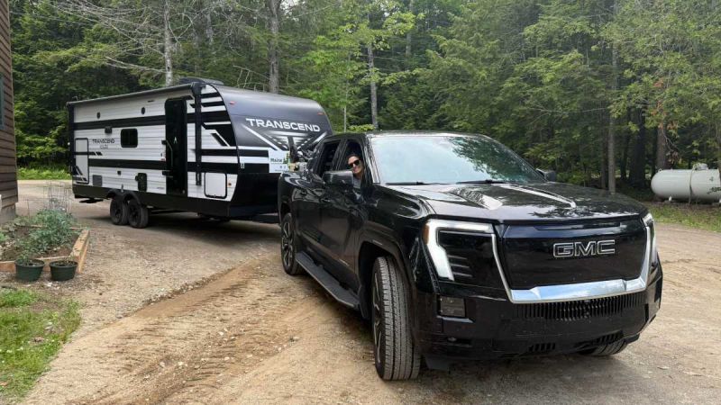 Black GMC Sierra EV towing white Transcend travel trailer through wooded campground with dense forest background