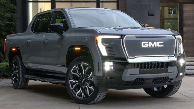 A sleek, silver GMC Sierra EV with illuminated headlights and grille parked outside a modern building, exuding a futuristic and robust design.