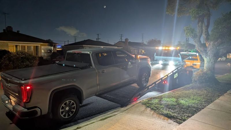 2025 GMC Sierra AT4 pickup truck in silver being loaded onto a tow truck at night in a residential neighborhood, illuminated by service lights.