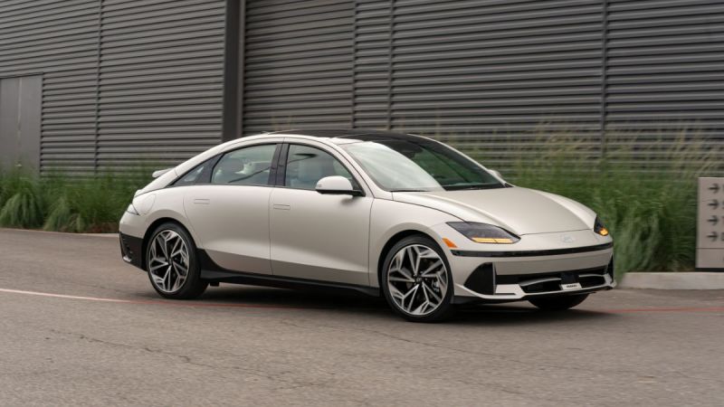 A white sedan parked on a street beside a corrugated metal building and greenery.