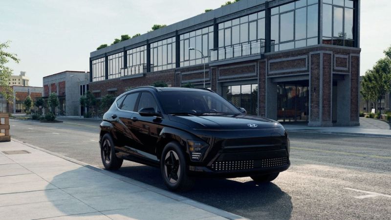 A black 2025 Hyundai Kona EV is parked on a quiet street beside a modern brick building with large glass windows. The setting is urban and calm, conveying elegance.