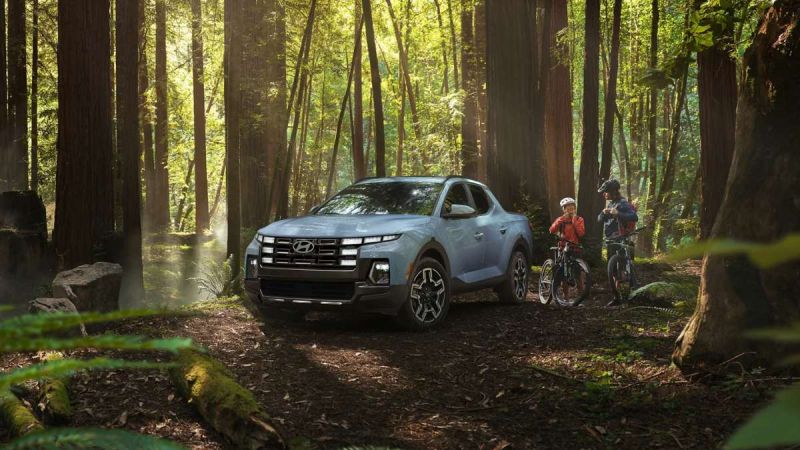 A silver Hyundai pickup truck is parked in a sunlit forest, surrounded by tall trees. Two cyclists in gear stand nearby, evoking adventure and exploration.