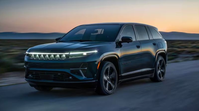 2025 Jeep Wagoneer in deep blue, driving on highway at sunset with mountain silhouette, LED headlights glowing
