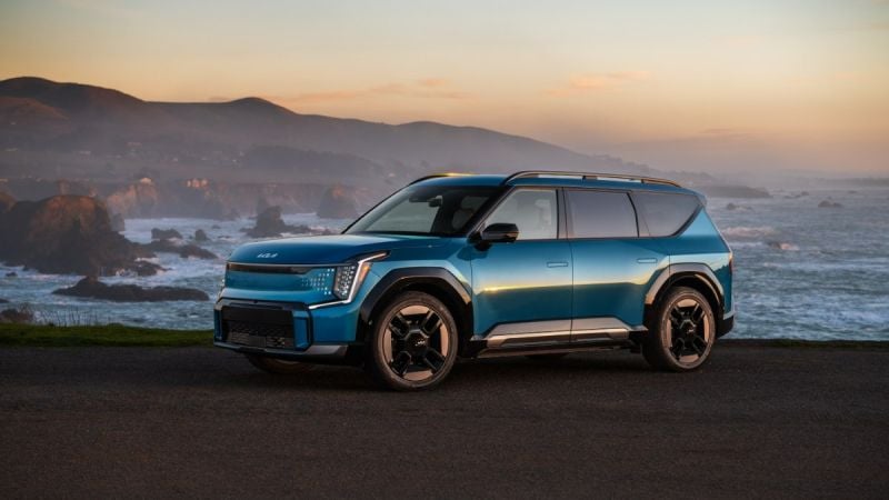 Blue 2025 Kia EV9 electric SUV parked near an ocean cliff at sunset, front three-quarter view.