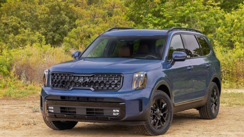 A blue 2025 Kia Telluride SUV shown from a front three-quarter angle, featuring its distinctive black grille and rugged off-road styling against a natural wooded background.