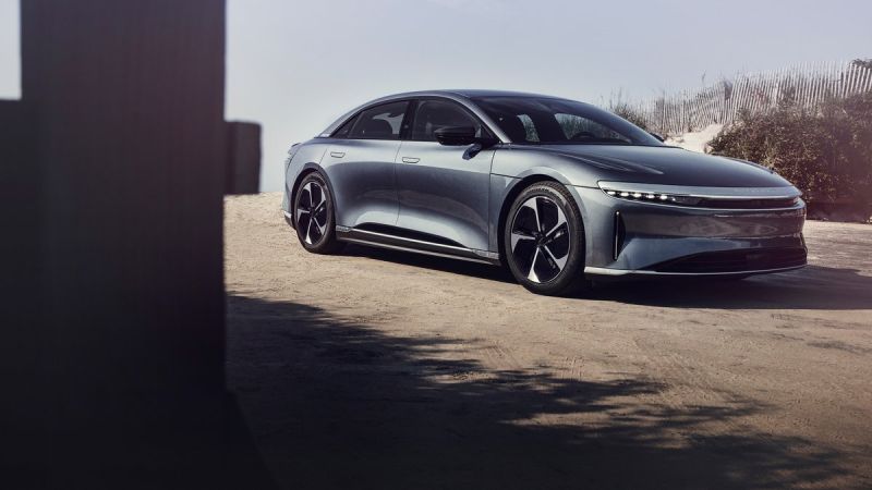 A gray 2025 Lucid Air Touring is parked on sandy terrain, framed by beach grass and wooden posts, showcasing its modern design.