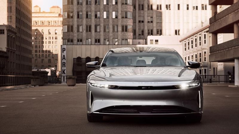 A silver 2025 Lucid Air parked in an urban setting, surrounded by tall buildings and a "PARK" sign in the background.